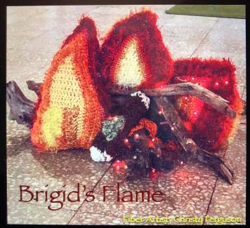 Picture of Yarnbomb installation named “Brigid’s Fire” by fiber artist and SIUE instructor, Christy Ferguson. The image shows a “campfire” made of three free-form crocheted and stuffed flames, a single crocheted and stuffed log complete with green moss accent, and Mississippi river driftwood. A single string of LED lights in red add to the glow of the crocheted representations of coals, ash, and embers that lie beneath.