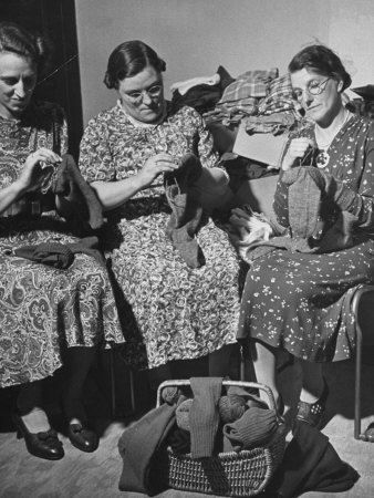 Black and white picture of three women engaged in a knitting circle or “stitch and bitch” during WWII. The women are sitting in chairs side by side, each wearing a calf-length dress in a different floral pattern. All three women are knitting what looks to be socks. The completed socks are piled in a wicker basket sitting on the floor in front of them.